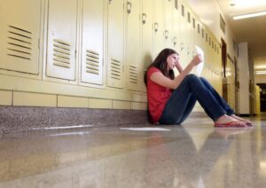A middle school girl sitting in hallway of school looks upset while looking at her classwork