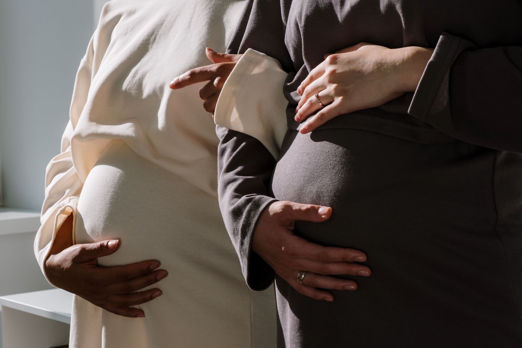two pregnant women standing side by side feeling their bellies